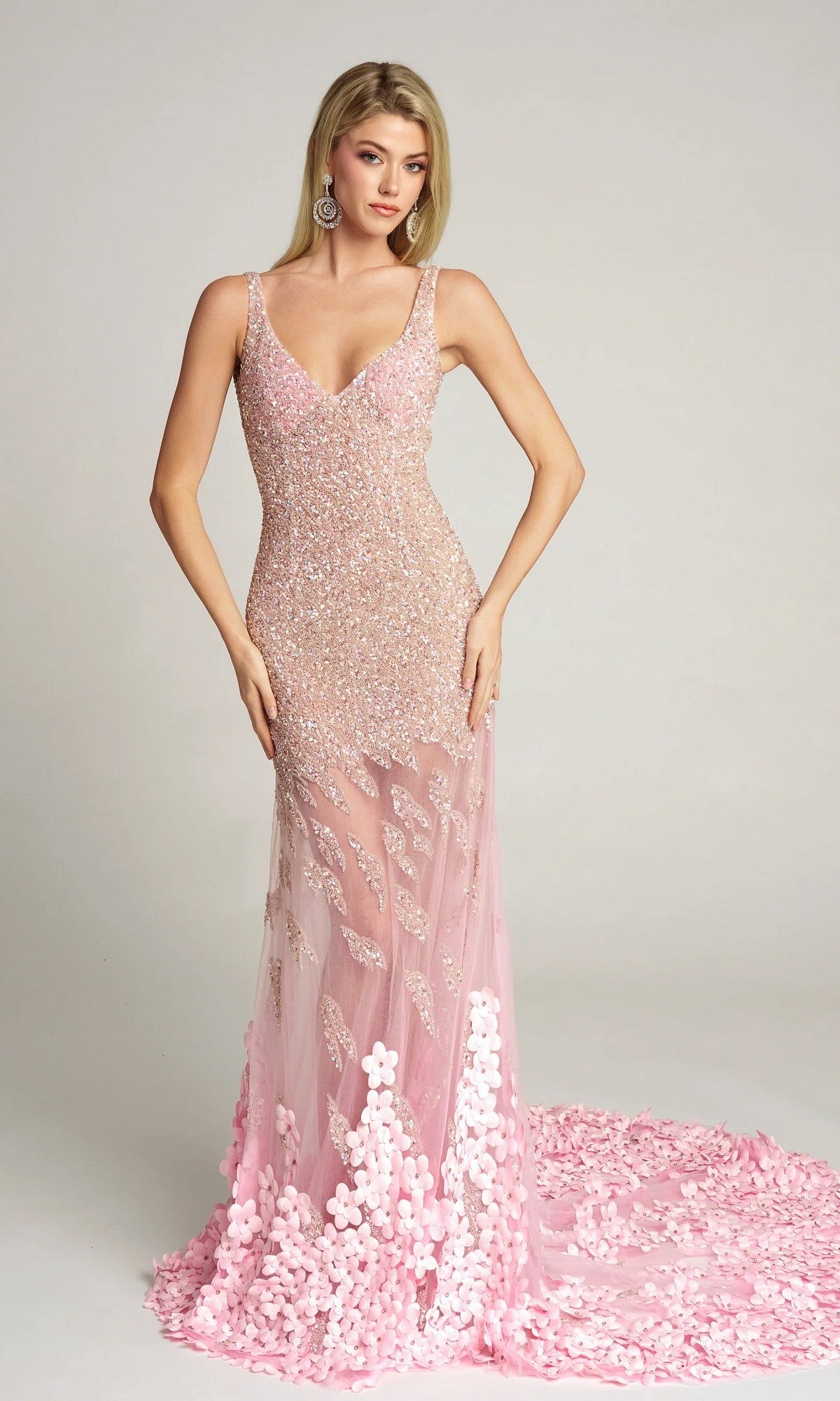 A woman wears the pink Portia and Scarlett Long Formal Dress (PS25987C), an embellished evening gown with sequin details, floral appliqués, and an illusion skirt. The sleeveless, floor-length dress contrasts with a plain light background.